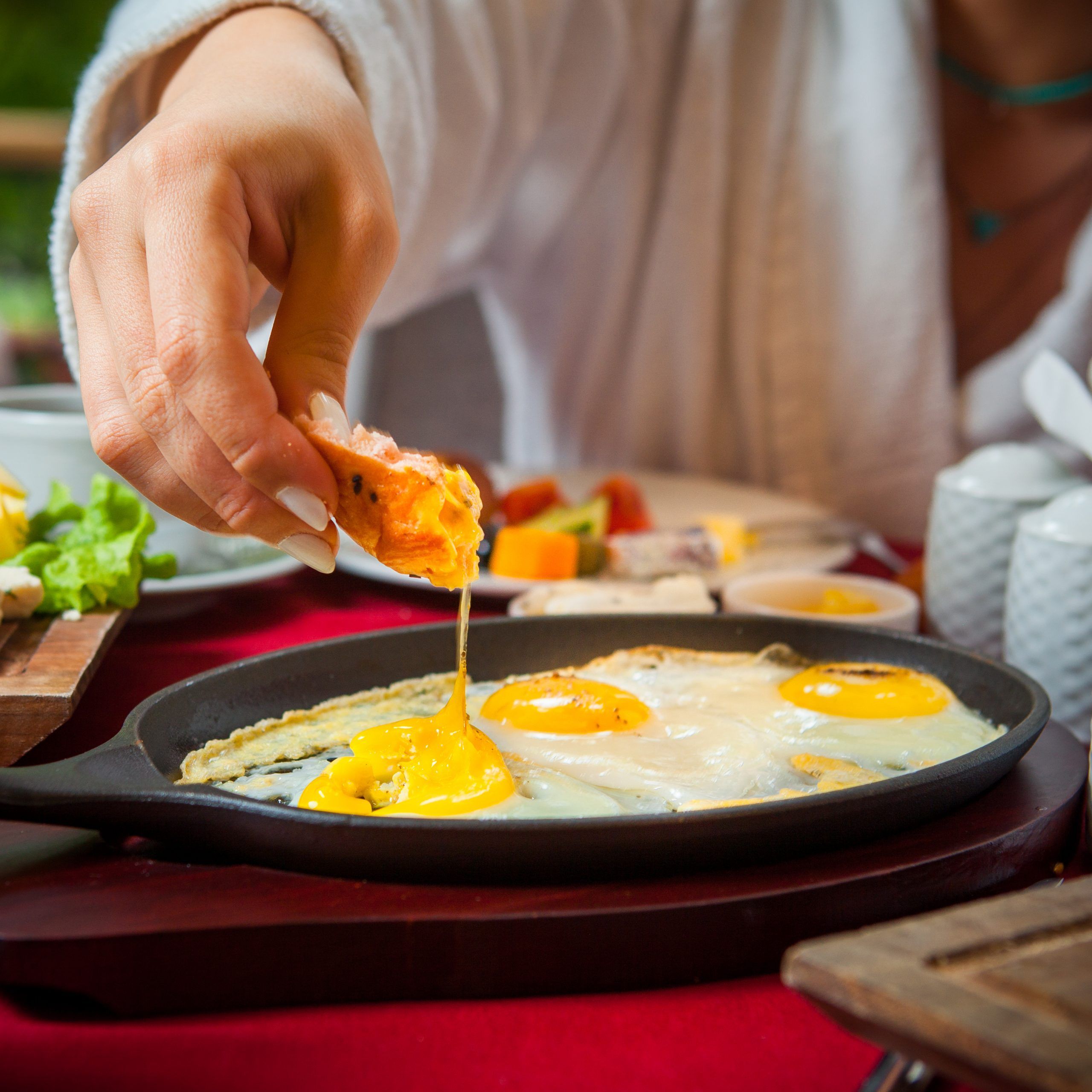 Close-up breakfast on a table with a red tablecloth fried eggs, cheese cheese, cucumbers, tomatoes, lettuce, coffee girl's hands